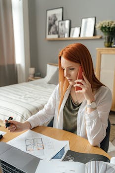 Redhead woman sits at a desk, focused on floor plans, using a smartphone, and working from home.